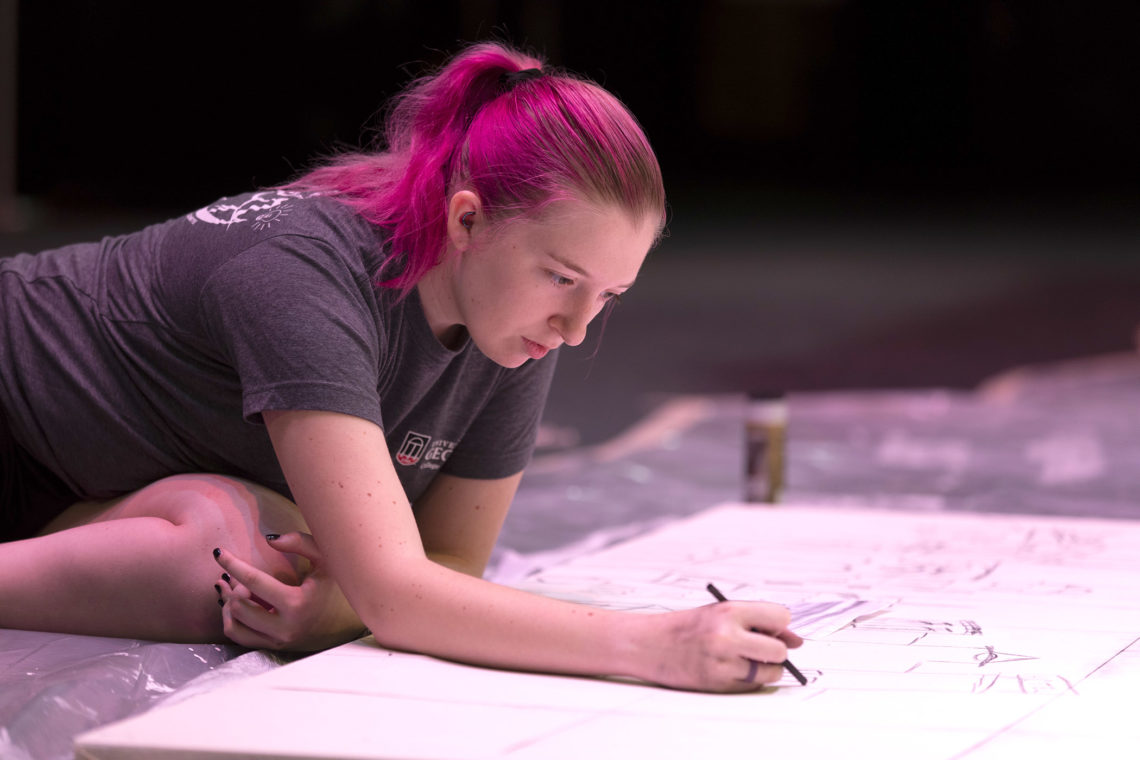 photo of woman leaning over a large drawing surface with pencils